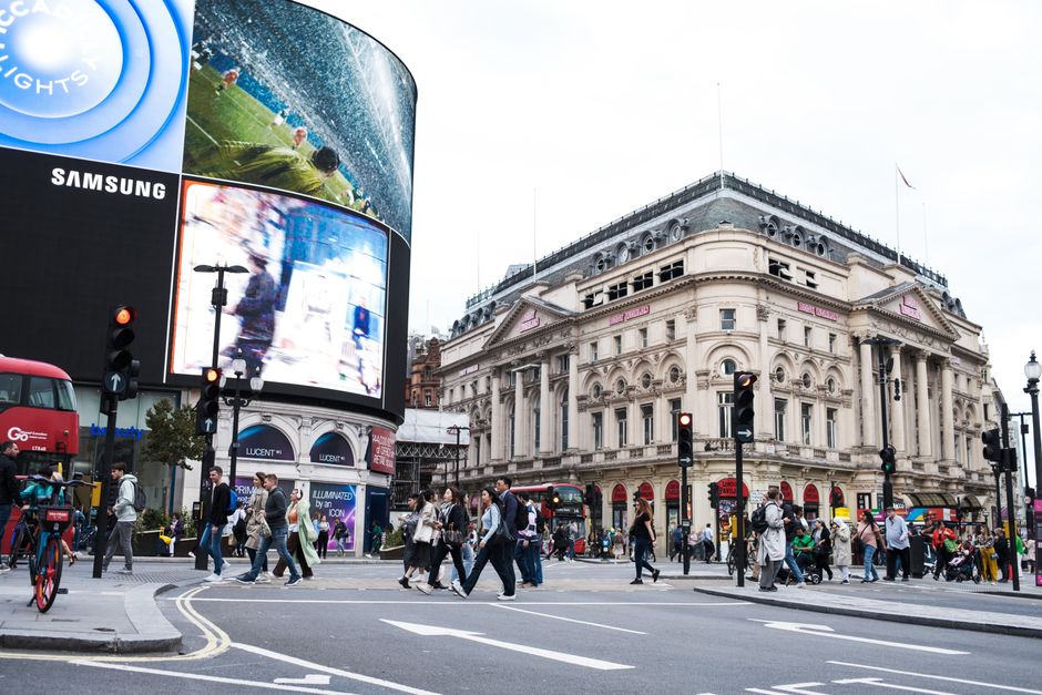 A nighttime street scene near Piccadilly Circus in central London, featuring a crowd of pedestrians gathering on the pavement and in front of illuminated buildings with ornate architectural details. P