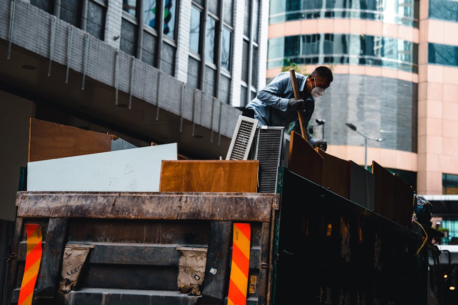 A worker wearing a white shirt, dark trousers, and a face mask is standing on the edge of a large, rusted industrial waste container mounted on a black truck bed, involved in sorting or handling bulky