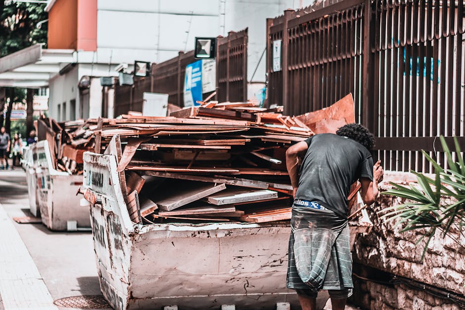 A worker wearing a white shirt, dark trousers, and a face mask is standing on the edge of a large, rusted industrial waste container mounted on a black truck bed, involved in sorting or handling bulky